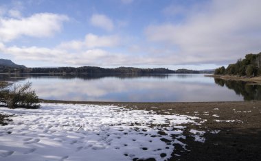 Patagonya 'daki büyülü kış manzarası. Kıyıdaki karın, gölün, dağların, ormanın ve mavi gökyüzünün beyaz bulutların yüzeydeki yansıması..