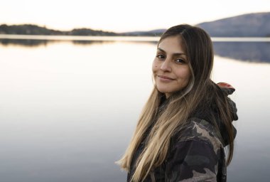 Gülümseyen genç bir kadının portresi. Arka planda durgun göl ve sıradağlar var. Güzel günbatımı renkleri. 