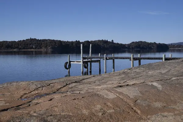 stock image The wooden dock in the placid lake in a summer sunny day. The rocky shore, forest and mountains in the background. 