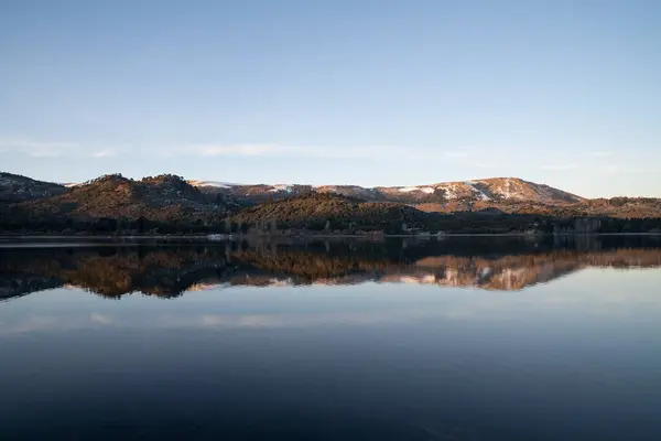 stock image The placid lake at sunset. The clear blue sky, forest and mountains in the horizon, reflected in the water surface. 