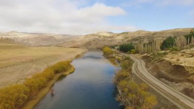 Kırsal alan. Sonbaharda sarı çayırlar boyunca akan nehrin yanında toprak yolun havadan görünüşü. Su yüzeyinde altın çayır, dağlar ve gökyüzü yansıması.