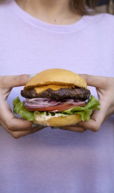 Hamburger sunumu. Yakından bakınca elinde marul, domates, çedar peyniri, kırmızı soğan ve mayonez olan bir kadın görülüyor.. 