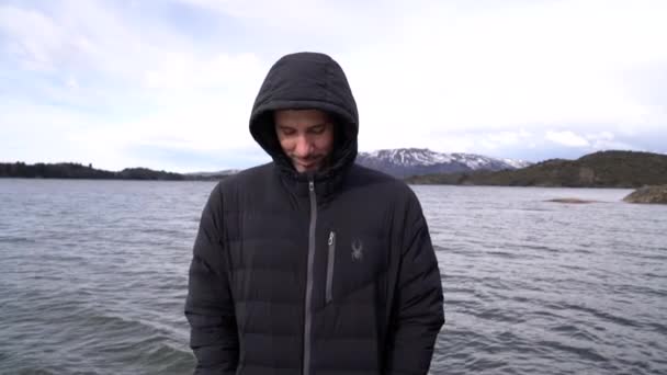 看到一个30多岁的白人穿着黑色夹克 看着相机 面带微笑 背景中的湖泊和山脉 — 图库视频影像