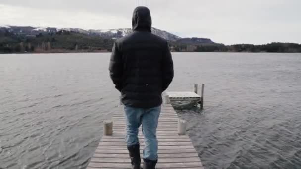 落ち着いた湖の木製ドックを歩いている黒いコートを着た男の姿 地平線の山と高山の森 — ストック動画