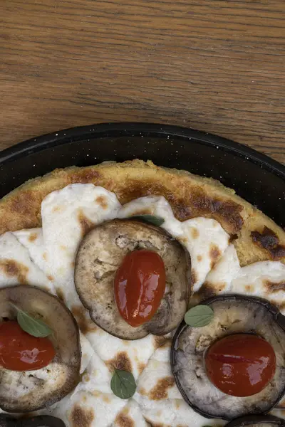 Vegan pizza. Top view of a vegetarian pizza with farinata, vegan mozzarella cheese, tomatoes, grilled eggplant and fresh oregano in a black pizza pan on the wooden table.