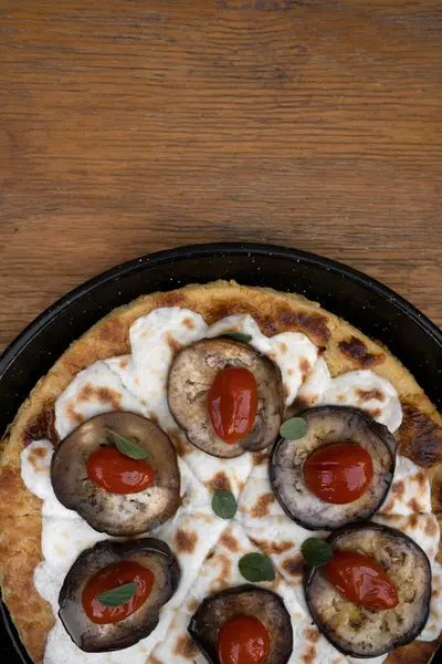 Vegan pizza. Top view of a vegetarian pizza with farinata, vegan mozzarella cheese, tomatoes, grilled eggplant and fresh oregano in a black pizza pan on the wooden table.