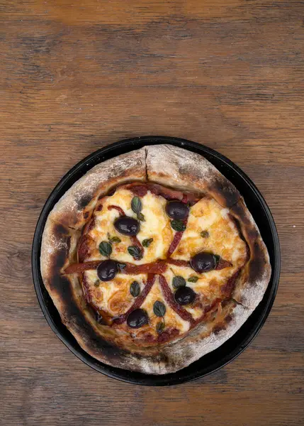 Pizza. Top view of a pizza with mozzarella cheese, grilled red bell peppers, ham, black olives and fresh oregano leaves, in a pizza pan on the wooden table.