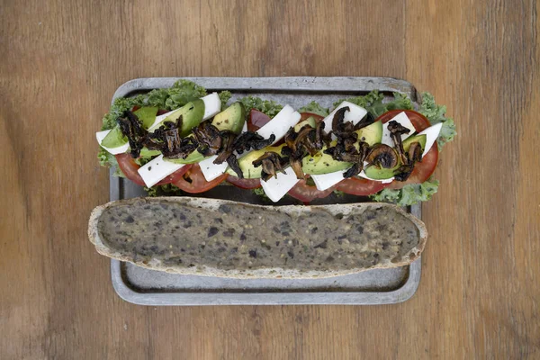 Vegan sandwich. Top view of a vegan baguette with tomato, vegan mozzarella, kale, avocado, babaganoush and mushroom chips, in a metal dish on the wooden table.
