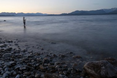 İyi geceler. Bulanık balıkçının karanlık silueti gece karanlığında gölde balık tutuyor. Kayalık kıyı, bulanık su ve ufukta güzel bir gün batımı ışığı olan dağlar..