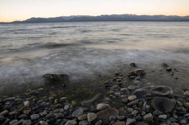 Gün batımında Nahuel Huapi Gölü 'nün uzun pozu. Güzel bulanık su efekti, kayalık sahil ve ufuktaki And Dağları sihirli bir alacakaranlık ışığıyla.