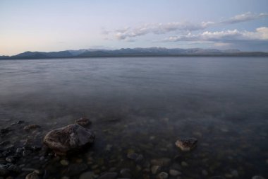 Gün batımında gölün gece görüntüsü. Kayalık kıyı manzarası, ufuktaki bulanık dalgalar ve dağlar, renkli bir alacakaranlık altında.. 