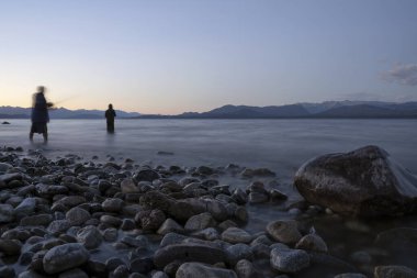 Uzun pozlu çekim. Gece karanlığında gölde balık tutan iki bulanık balıkçının karanlık siluetinin görüntüsü. Kayalık kıyı, bulanık su ve ufukta alacakaranlık ışığı olan dağlar..