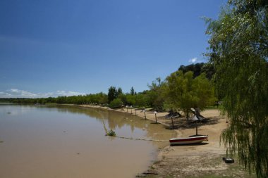 Güneşli bir yaz gününde nehir kıyısı manzarası. Arka plandaki tropikal ormandaki kumda bir tekne suya yansıyor..