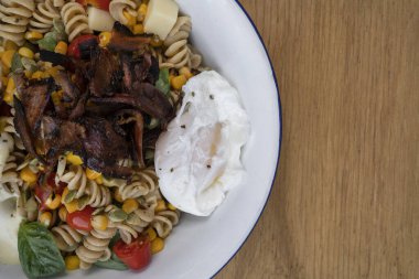 Fusilli makarna salatasının üst görüntüsü fesleğen, kıtır pastırma, mısır, peynir, domates ve ahşap restoran masasının üzerinde kaynatılmış yumurta..