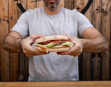 Yakından bakınca elinde et, marul, mozzarella peyniri ve domatesli sandviç tutan bir adam görülüyor..