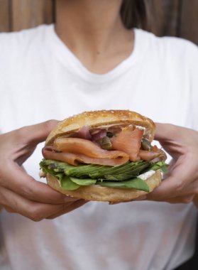 Yemek yiyorum. Yakından bakınca, elinde somonlu sandviç tutan bir kadın görülüyor. Lezzetli simit katmanları, pembe somon fileto, krem peynir, dilimlenmiş avokado, kapari, soğan ve roka..