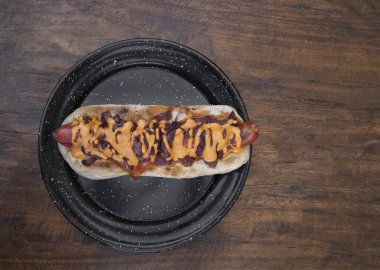 Lezzetli bir sosisli sandviçin üst görüntüsü. Çıtır pastırma, soğan ve çedar peyniri. Lokanta tahtasının üstünde siyah bir tabakta..