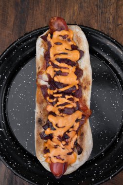 Lezzetli bir sosisli sandviçin üst görüntüsü. Çıtır pastırma, soğan ve çedar peyniri. Lokanta tahtasının üstünde siyah bir tabakta..