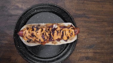 Lezzetli bir sosisli sandviçin üst görüntüsü. Çıtır pastırma, soğan ve çedar peyniri. Lokanta tahtasının üstünde siyah bir tabakta..