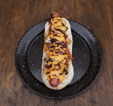 Lezzetli bir sosisli sandviçin üst görüntüsü. Çıtır pastırma, soğan ve çedar peyniri. Lokanta tahtasının üstünde siyah bir tabakta..