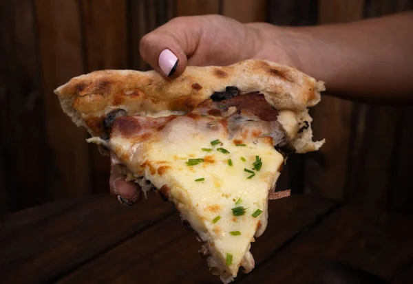 Eating pizza. Female hand holding a slice of pizza with mozzarella cheese, mushrooms, crispy bacon, sliced chives and garlic, on the restaurant wooden table.