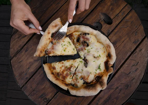 Eating pizza at the restaurant. Top view of female hands serving a slice of pizza with mozzarella cheese, mushrooms, bacon, sliced chives and garlic.