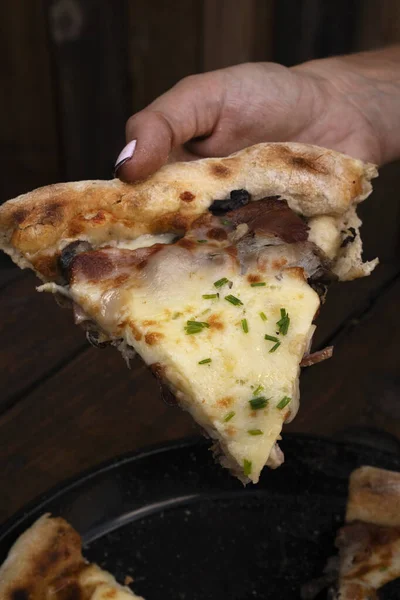 Eating pizza. Female hand holding a slice of pizza with mozzarella cheese, mushrooms, crispy bacon, sliced chives and garlic, on the restaurant wooden table.