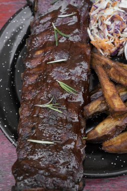 Barbekü sosu ve otla marine edilmiş domuz pirzolası, tatlı patates kızartması ve lahana salatası, masada siyah bir tabakta..