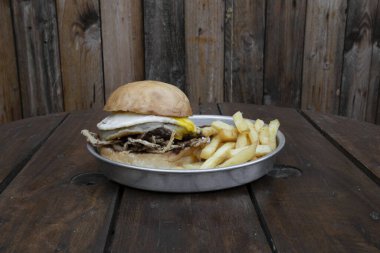 Amerikan yemekleri kırsal sunumu. Et, ızgara yumurta, kızarmış soğan, provolone peyniri, patates ekmeği ve patates kızartması ile ahşap bir tabakta lezzetli bir hamburgerin yakın görüntüsü.. 