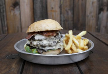 Canavar burger. Eti, marulu, domatesi, peynirli pastırması ve patates kızartması olan dev bir hamburgerin yakından görüntüsü, ahşap bir tabakta servis ediliyor.. 