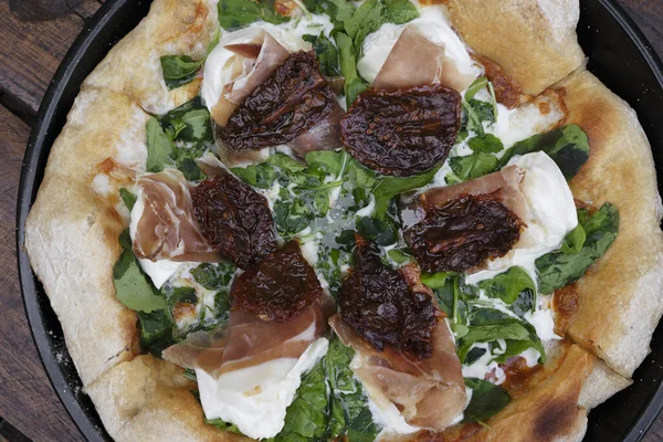 Burrata peynirli roka pizzası, kurutulmuş domates ve çiğ jambon, ahşap masada pizza tavasında servis edilir.. 