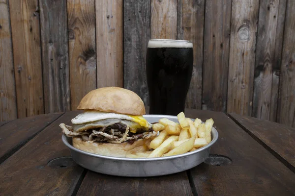 Restaurant menu. Closeup view of a burger with meat, cheese, onions and a grilled egg, with french fries and a dark beer.