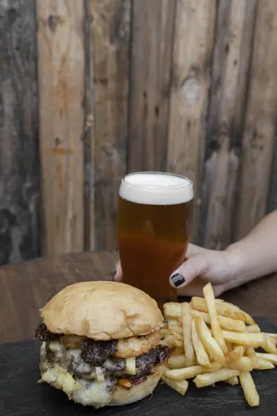 Eating at the pub. Delicious rustic burger with beef, fried onion rings, brie cheese, almonds, sun dried tomatoes and sweet chili, with french fries. A woman hand holding a fresh glass of beer.