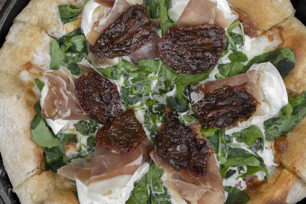 Burrata peynirli roka pizzası, kurutulmuş domates ve çiğ jambon, ahşap masada pizza tavasında servis edilir.. 