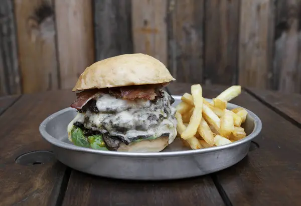 Monster burger. Closeup view of a giant burger with meat, lettuce, tomato, cheese bacon and french fries, served on a metal plate on the wooden table.