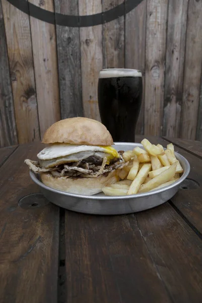 Restaurant menu. Closeup view of a burger with meat, cheese, onions and a grilled egg, with french fries and a dark beer.