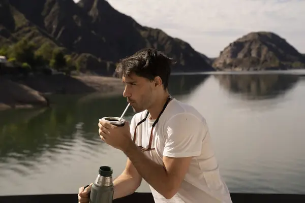 Drinking mate in the morning. Closeup view of a young man drinking traditional yerba mate herb infusion drink, with the lake and mountains in the background.
