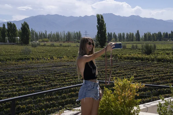 Travel. View of a young woman taking a selfie with her cellphone in the vineyard. The grapevines plantation and mountains in the background.