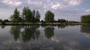 Gölün ve golf sahasının manzarası. Gölet turu, kıyı şeridi ve çim alanındaki ağaçlar. Su yüzeyindeki ağaçların ve gökyüzünün güzel yansıması..