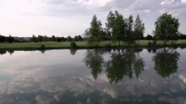 Gölün ve golf sahasının manzarası. Gölet turu, kıyı şeridi ve çim alanındaki ağaçlar. Su yüzeyindeki ağaçların ve gökyüzünün güzel yansıması..