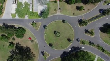 Overhead aerial view of a roundabout in a residential neighborhood in the suburbs. We can see a few cars passing by, a tennis court and houses.