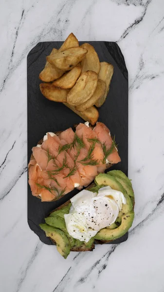Healthy cuisine. Gourmet sandwich. Top view of a smoked salmon, cream cheese, sliced avocado and poached eggs sandwich with fried potatoes on a black dish on the white marble table.