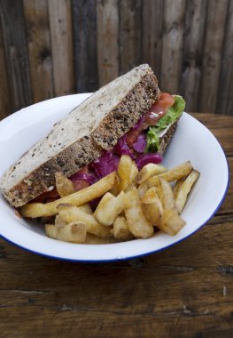 Ekmek, tütsülenmiş somon, krem peynir, roka, mor soğan ve garnitür olarak patates kızartmasından yapılmış bir sandviç. Restoranın ahşap masasında beyaz bir kasede.. 