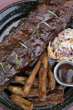 Barbekü sosu ve otla marine edilmiş domuz pirzolası, tatlı patates kızartması ve lahana salatası, masada siyah bir tabakta..
