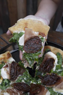 Yakından bakınca, elinde burrata peyniri, güneşte kurutulmuş domates ve çiğ jambon olan bir dilim pizza tutan bir kadın eli görülüyor..