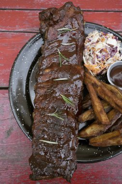 Barbekü sosu ve otla marine edilmiş domuz pirzolası, tatlı patates kızartması ve lahana salatası, masada siyah bir tabakta..