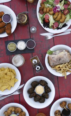 Restoranın ahşap masasında farklı yemekler, cipsler, börekler, sandviç, patates kızartması, ekmek ve salata..