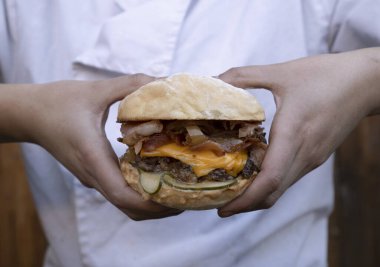 Kafkasyalı kadın şef elleriyle canavar burger tutuyor. Ekmek, et, çedar peyniri, salatalık turşusu, pastırma ve sos katmanları.. 