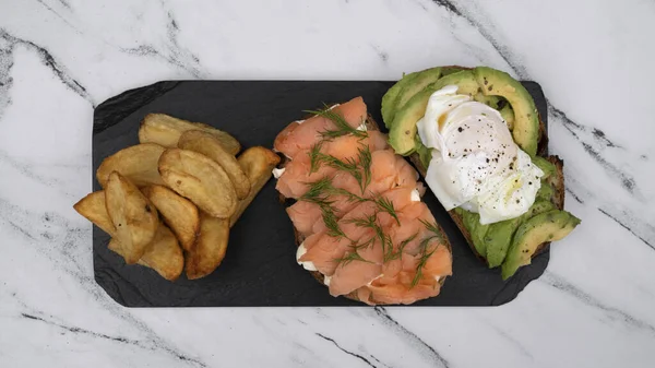 Healthy cuisine. Gourmet sandwich. Top view of a smoked salmon, cream cheese, sliced avocado and poached eggs sandwich with fried potatoes on a black dish on the white marble table.
