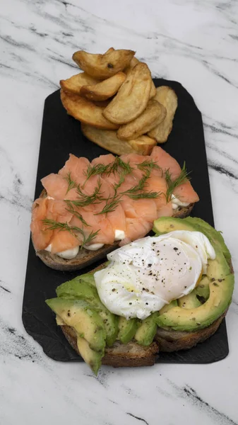 Healthy cuisine. Gourmet sandwich. Closeup view of a smoked salmon, cream cheese, sliced avocado and poached eggs sandwich with fried potatoes on a black dish on the white marble table.